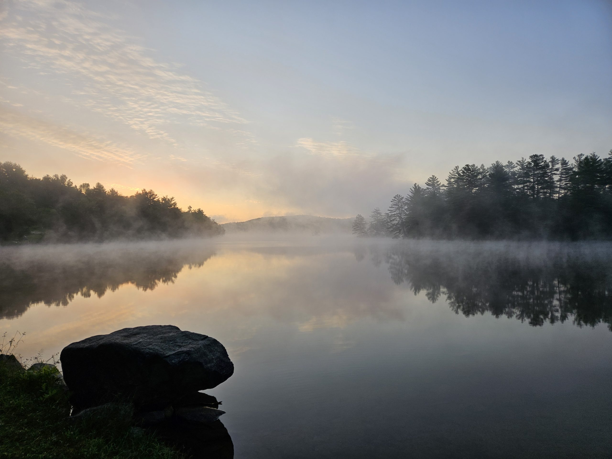 A large boulder at the edge of a still pond at dawn, mist rising, trees reflected in the water