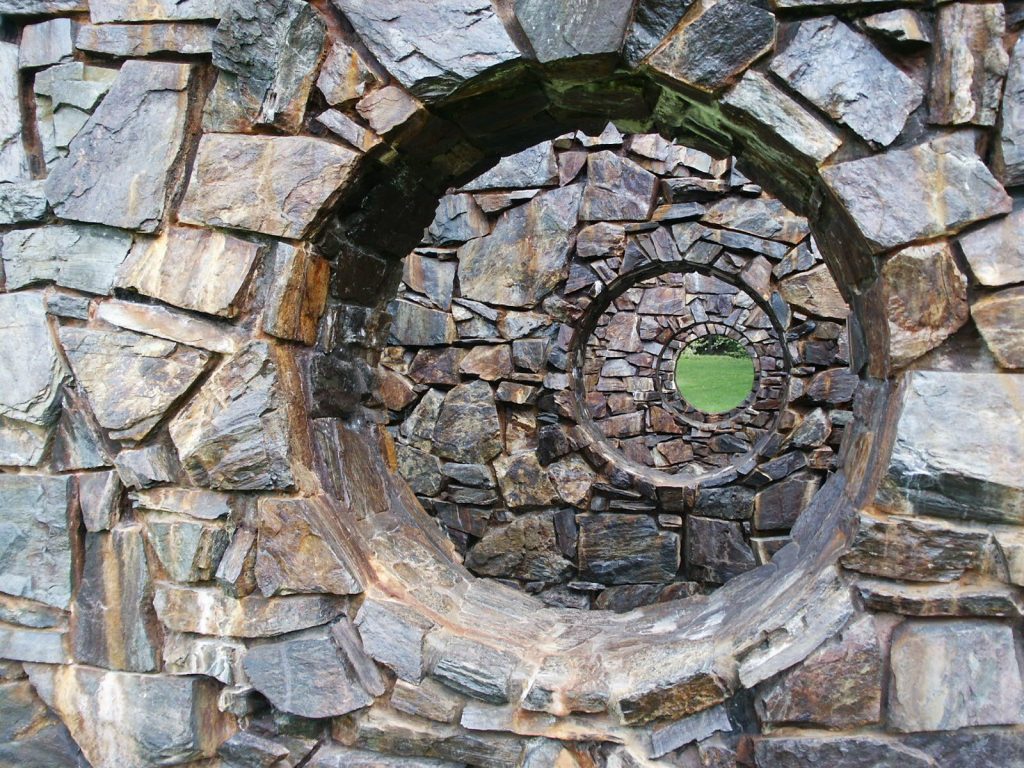Intricate spiral of nested circular openings carved through a stone wall, with a glimpse of green grass visible at the center