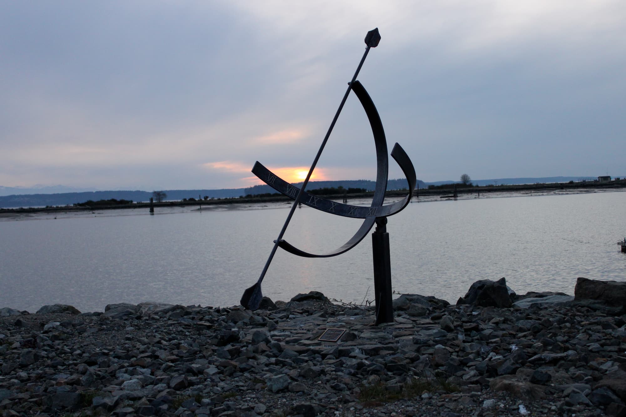 A sculptural iron sundial with Roman numerals stands on a rocky shoreline at sunset, water and distant hills behind it, the setting sun glowing through overcast clouds.